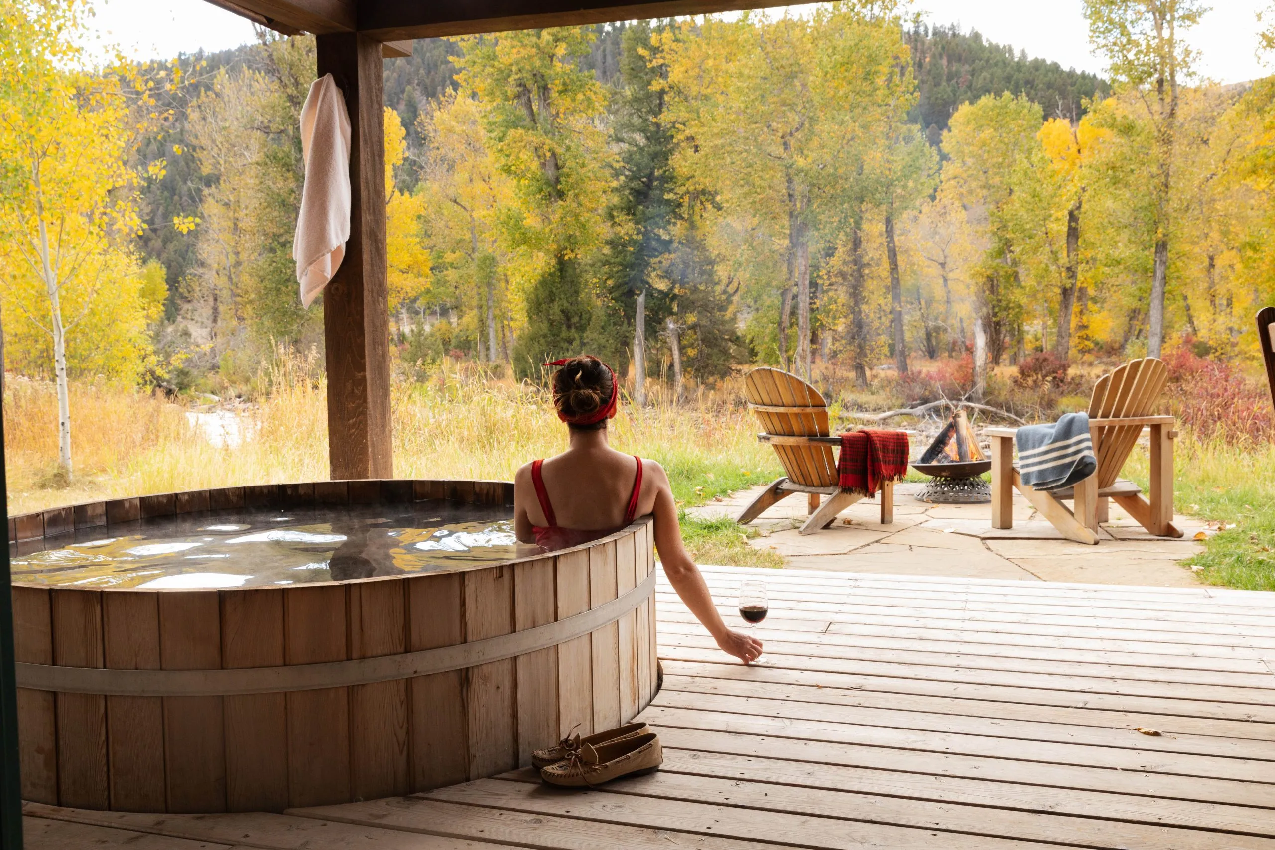 A woman relaxing while holding a glass of red wine sitting inside a wooden hot tub on a porch next to a fire pit and lounge chairs