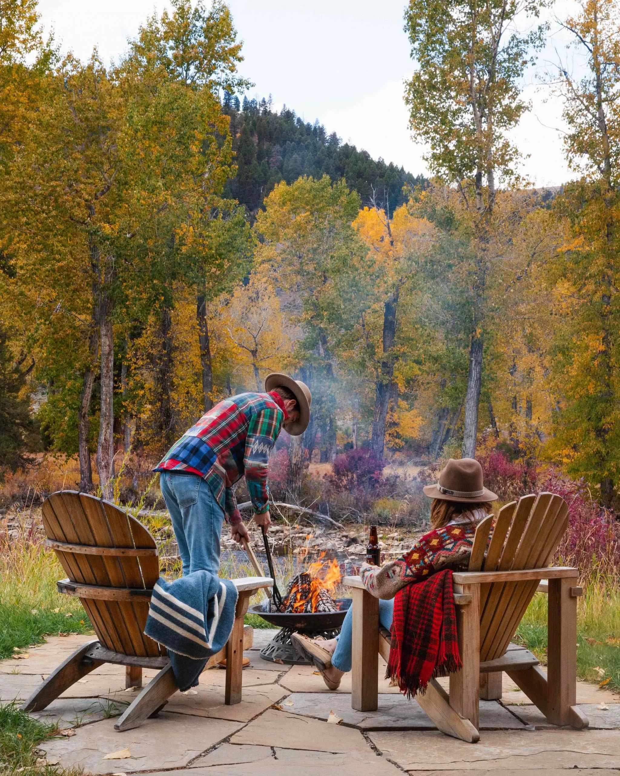 A woman sitting in a wooden chair holding a beer while a man tends to a fire pit