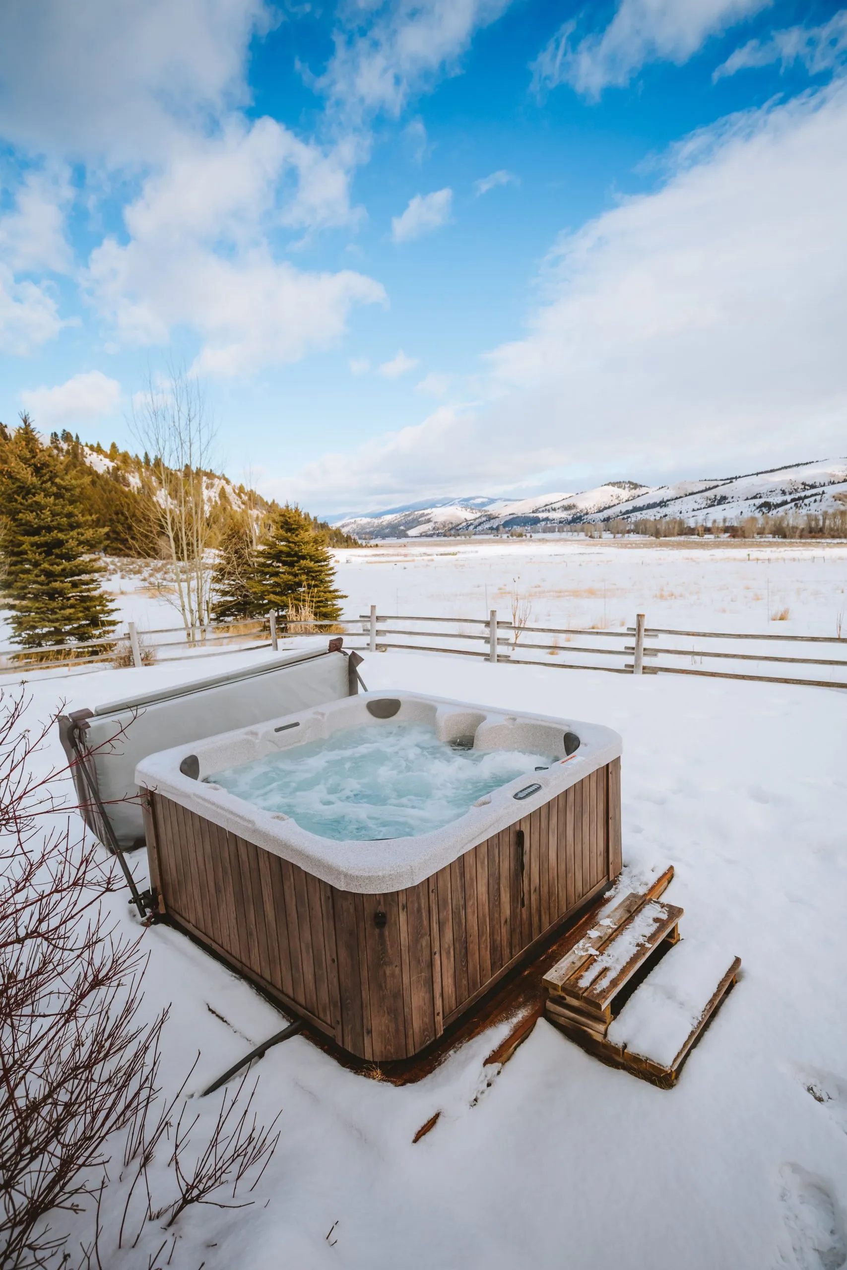 A hot tub outside on a wooden platform with snow covering the ground, blue sky and clouds in the distance