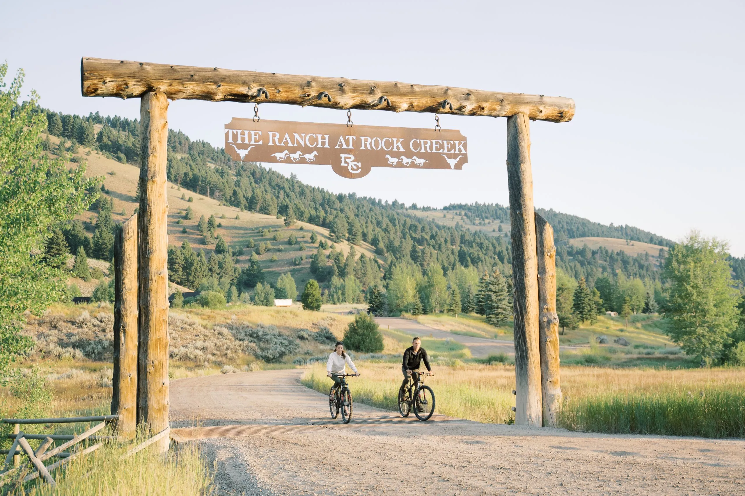 A couple riding mountain bikes under an arch with a sign that reads "The Ranch at Rock Creek"