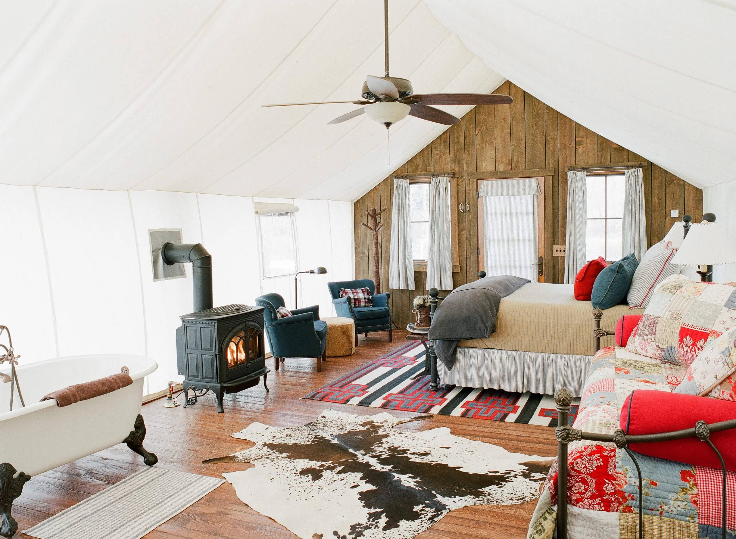 A bedroom inside a tent cabin with a hanging ceiling fan, wood burning fireplace, bathtub, animal skin rug, and a bench with a quilt with matching pillows