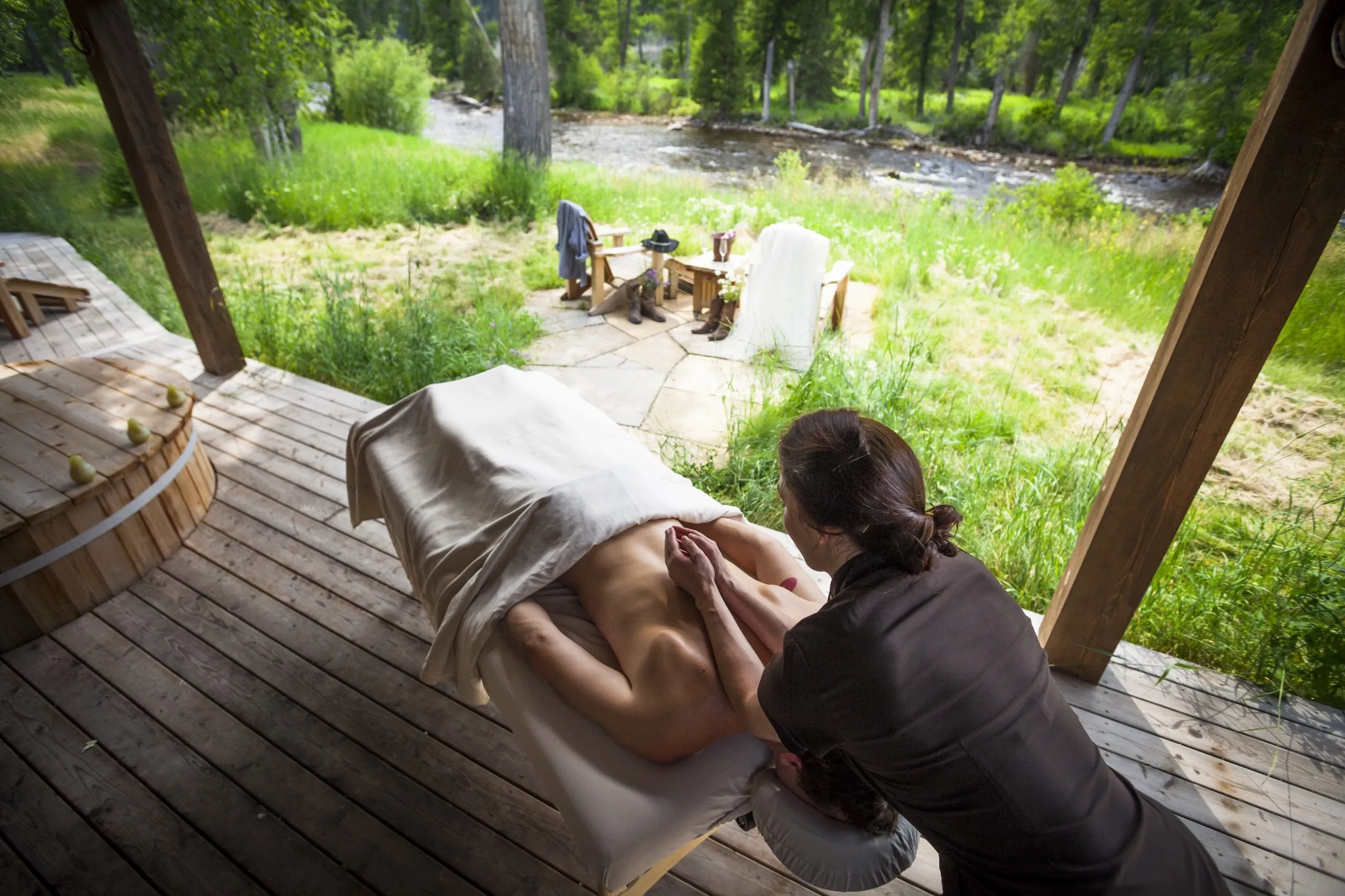 A masseuse massaging a client on a massage table located on a scenic porch facing a river